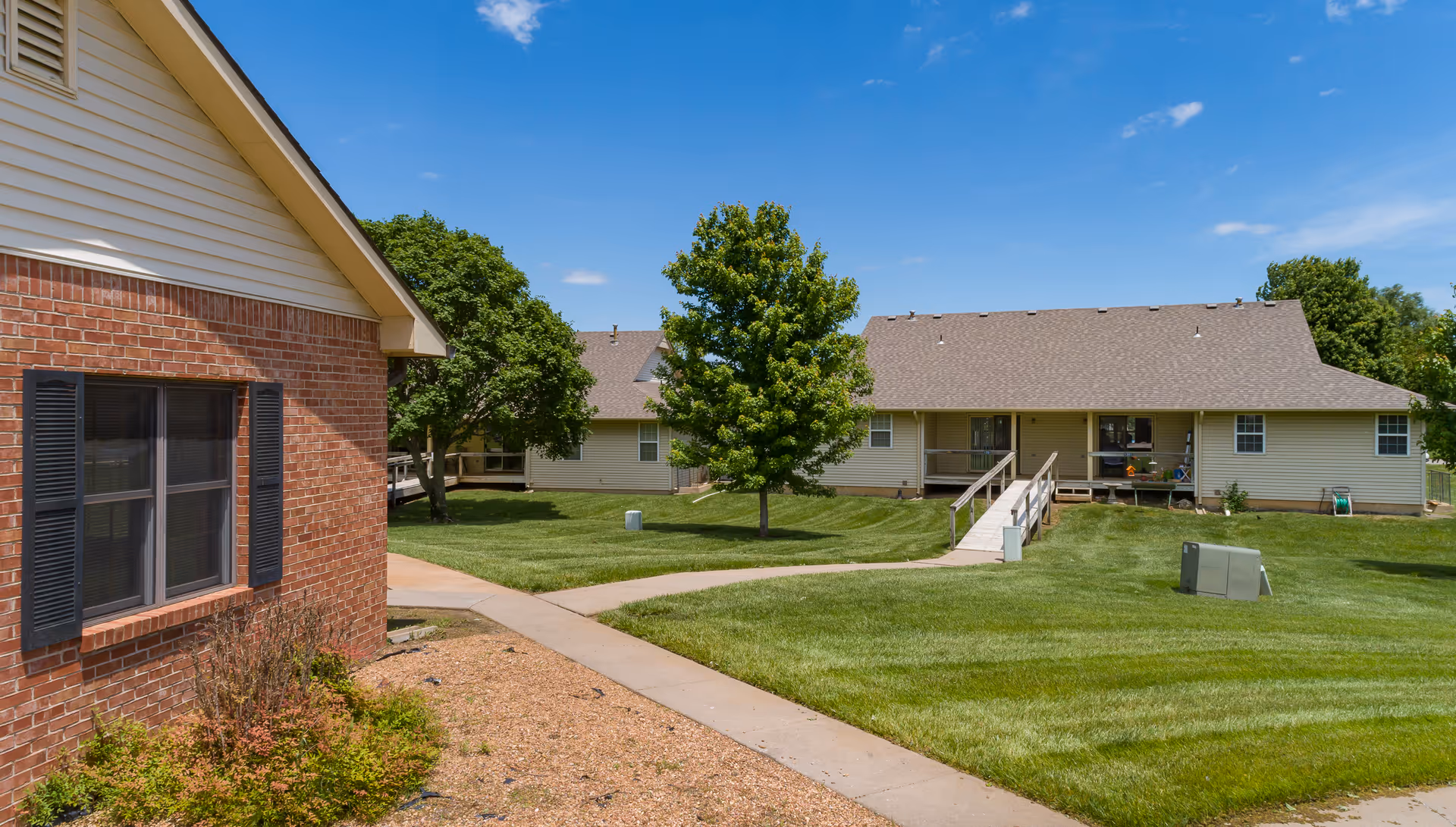 View of a senior living facility exterior with a brick building on the left and a beige building with a ramp on the right, surrounded by green grass, trees, and a clear blue sky.