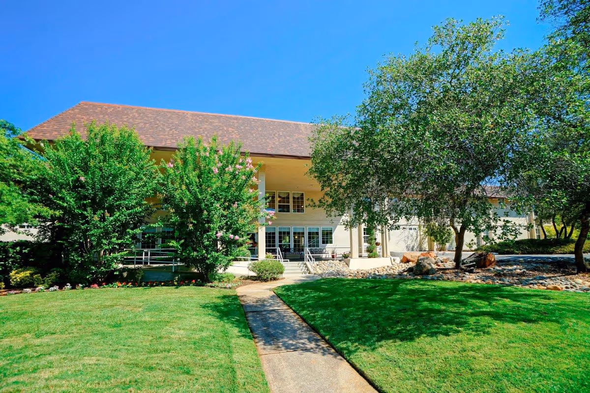 Exterior view of Eskaton Cameron Park facility showing a two-story building with a brown roof, surrounded by green trees and a well-maintained lawn under a clear blue sky. A concrete pathway leads to the entrance with steps and railings.