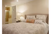 A cozy bedroom with a neatly made bed featuring patterned bedding and multiple pillows. A bedside table with a lamp is next to the bed. An open door reveals a bathroom with a shower stall.