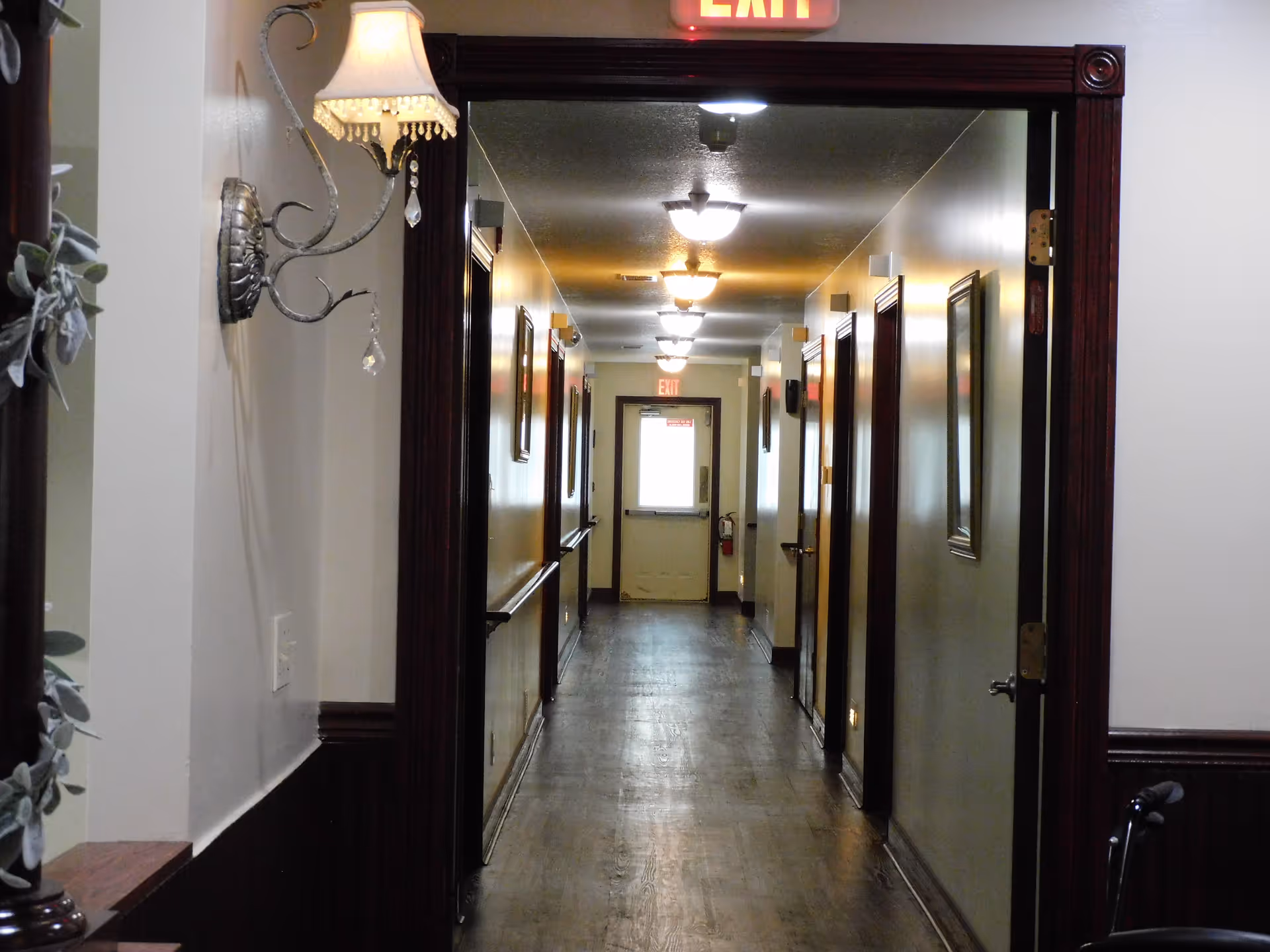 A long, narrow hallway in an assisted living facility with wooden floors and cream-colored walls. The hallway is lined with several closed doors on both sides, framed pictures on the walls, and ceiling lights evenly spaced along the length. There is an exit sign above a door at the far end of the hallway. A decorative wall sconce with a lampshade is visible on the left side near the foreground.