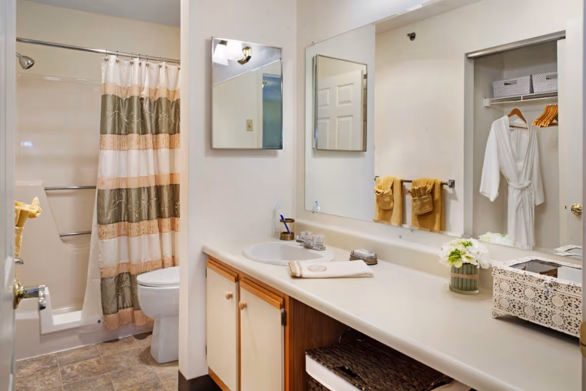 A bathroom with a shower and toilet on the left side, a striped shower curtain, a large mirror above a countertop with a sink, towels hanging on a rack, a white robe hanging in an open closet, and decorative items on the counter including a small vase with flowers and a decorative box.