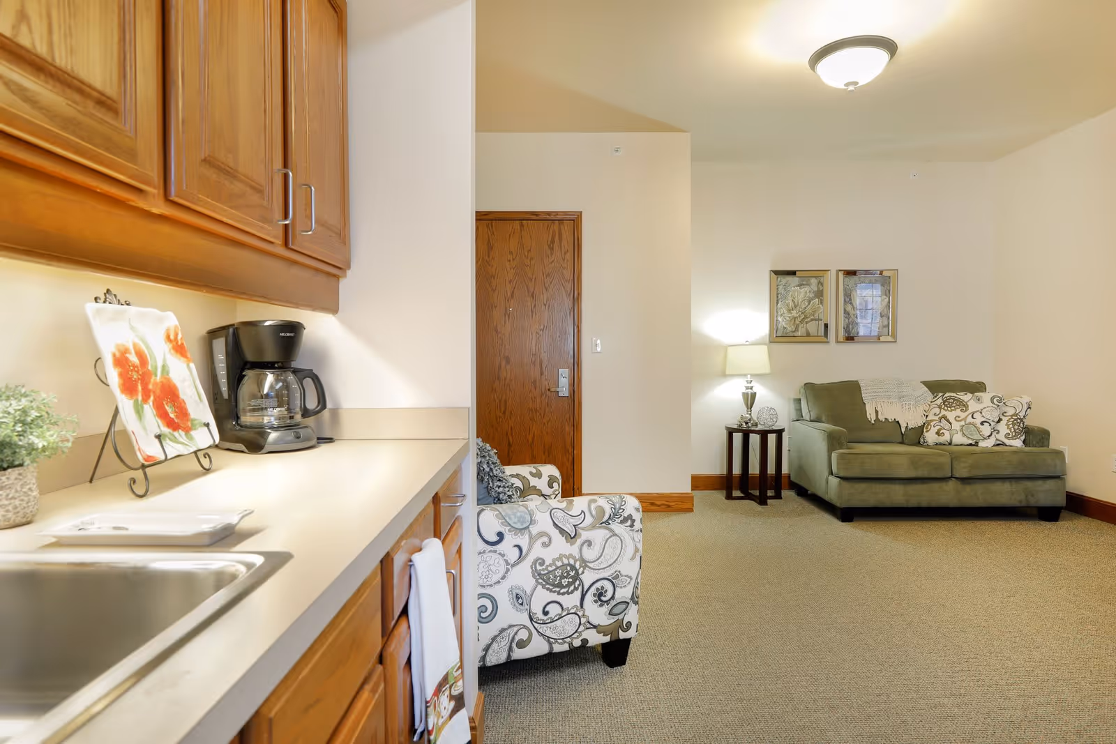 Interior view of a senior living facility room showing a small kitchenette area with wooden cabinets, a coffee maker, and a sink on the left side. In the background, there is a seating area with a patterned armchair, a green sofa with decorative pillows, a side table with a lamp, and two framed pictures on the wall. The room has beige walls and carpeted flooring.