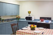 Small institutional-style kitchen with blue cabinets, a table covered by a red-and-white checkered tablecloth set for four, a microwave and stove, and fruit bowls.