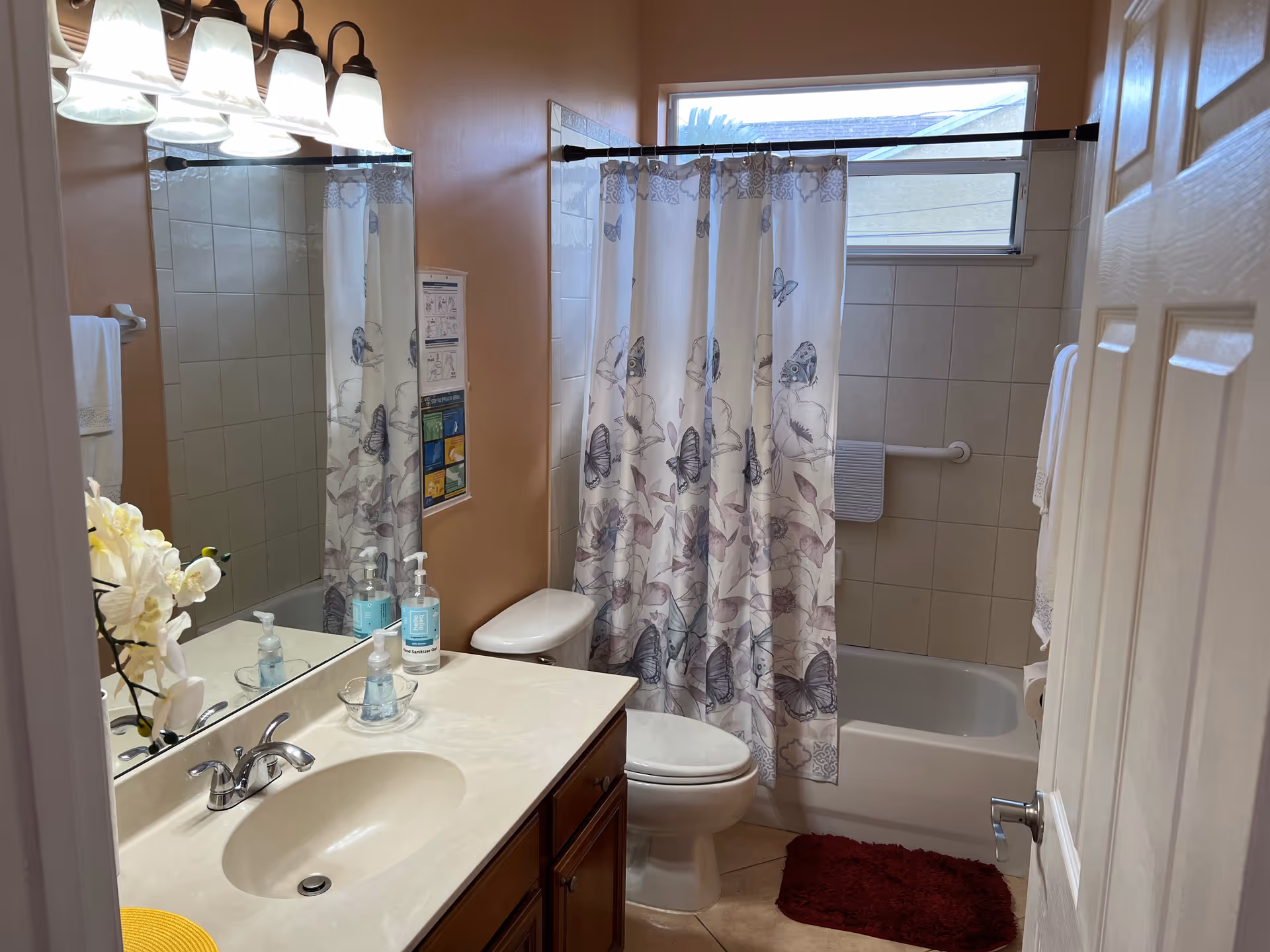 A clean bathroom with a single sink vanity, a large mirror, and a light fixture with five bulbs above it. There is a white toilet next to a bathtub with a shower curtain featuring a butterfly pattern. A window above the bathtub allows natural light in. The walls are painted a warm beige color, and there are handrails installed near the bathtub for safety. A red bath mat is placed on the tiled floor.