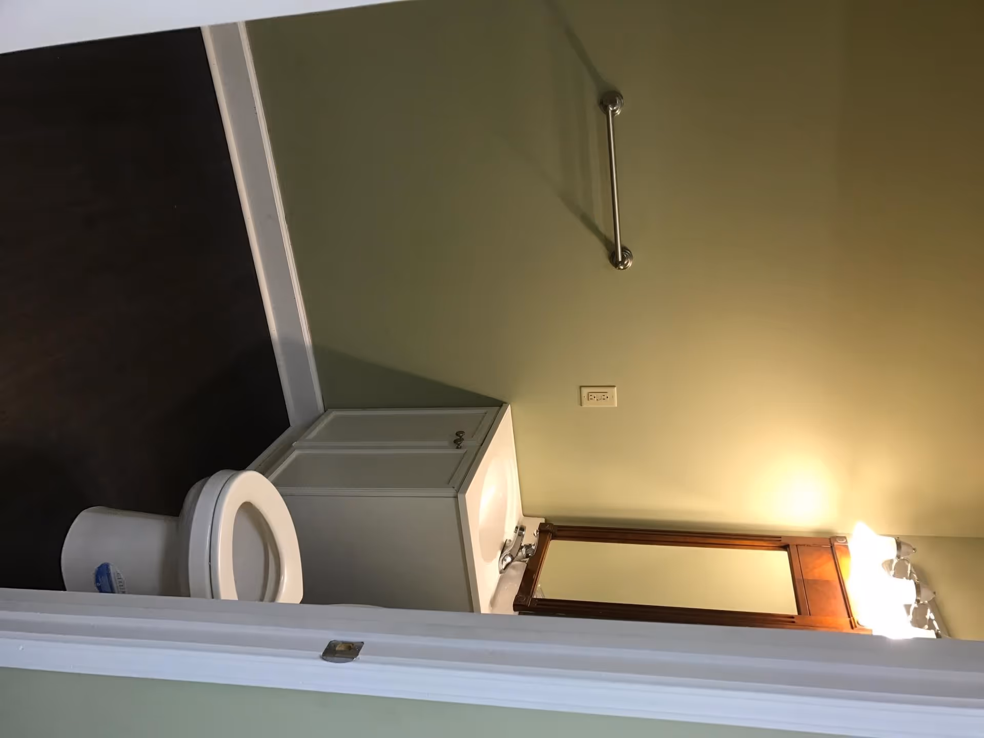 A small bathroom with a white toilet, a white vanity with a sink, a wooden framed mirror above the sink, a light fixture with three bulbs above the mirror, and a metal grab bar mounted on the light green wall.