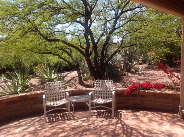 Two woven patio chairs and a small table on a shaded tiled patio with desert landscaping, trees, agave plants, and red flowering shrubs.