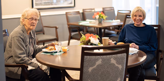 Two elderly women sitting at a round wooden table in a dining area, enjoying a meal with plates, glasses, and a floral centerpiece on the table. The room has several chairs and tables, with soft lighting and a framed picture on the wall.