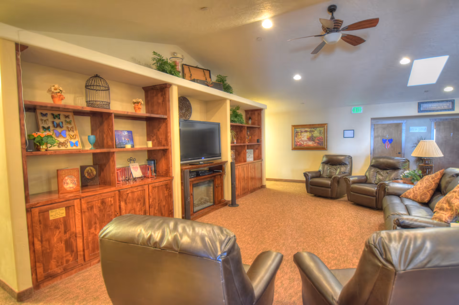 A cozy living room area in a senior living facility featuring brown leather armchairs and a sofa arranged around a TV mounted on a wooden entertainment center with shelves displaying decorative items. The room has warm lighting, a ceiling fan, carpeted floor, and a framed painting on the wall.