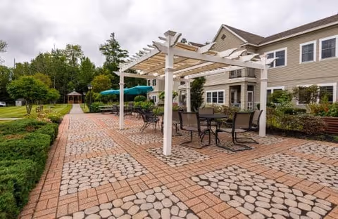Paved outdoor patio at Granite Hill Estates with a white pergola, tables and chairs, landscaped grounds and a distant gazebo beside the building.