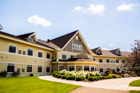 Exterior view of a large, two-story senior living facility building with beige and yellow siding, multiple windows, and a peaked roof. The foreground features a well-maintained lawn, a curved walkway, and landscaped bushes with white flowers under a partly cloudy blue sky.