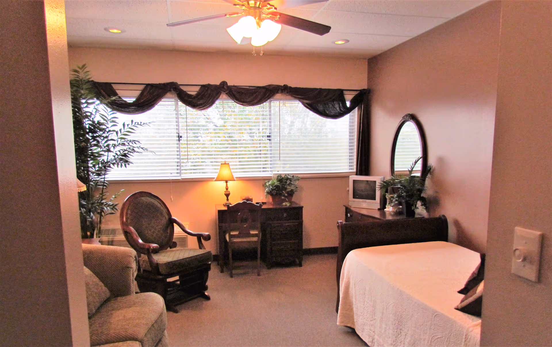 A cozy bedroom with a single bed covered in a white quilt, a wooden dresser with a small TV and plants on top, a round mirror on the wall, a wooden desk with a chair and a lamp, a cushioned armchair, and a large window with blinds and dark curtains. There is also a potted plant in the corner and a ceiling fan with lights.
