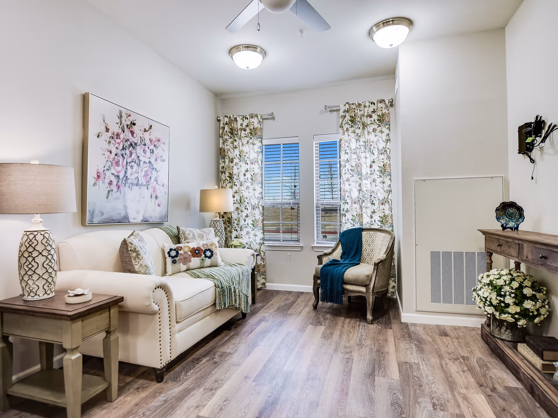 A cozy living room with a white sofa adorned with decorative pillows and a green throw blanket. Next to the sofa is a wooden side table with a patterned lamp. Two windows with floral curtains let in natural light. An armchair with a blue throw is positioned near the windows. A wooden console table with decorative items and a potted plant is against the wall. The room has wood flooring and white walls with ceiling lights and a ceiling fan.