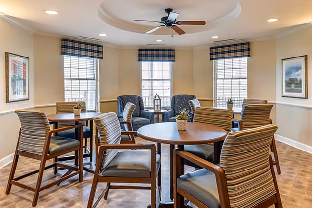 A bright and cozy common area with round wooden tables surrounded by striped cushioned chairs. Three large windows with plaid valances allow natural light to fill the room. Two blue upholstered armchairs and a small table with a decorative lantern are positioned near the windows. The ceiling features a recessed circular design with a ceiling fan in the center. Framed artwork hangs on the beige walls.