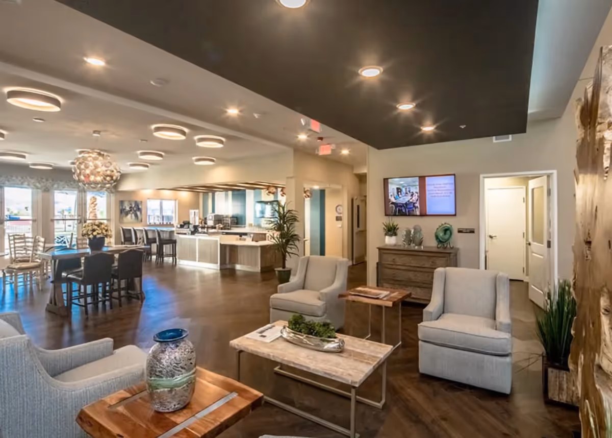 A modern and spacious interior common area with comfortable armchairs, wooden tables, a decorative vase, and a chest of drawers with plants and decorative items. The room features a dark ceiling with recessed lighting, a wall-mounted TV, and a dining area with tables and chairs near large windows letting in natural light.