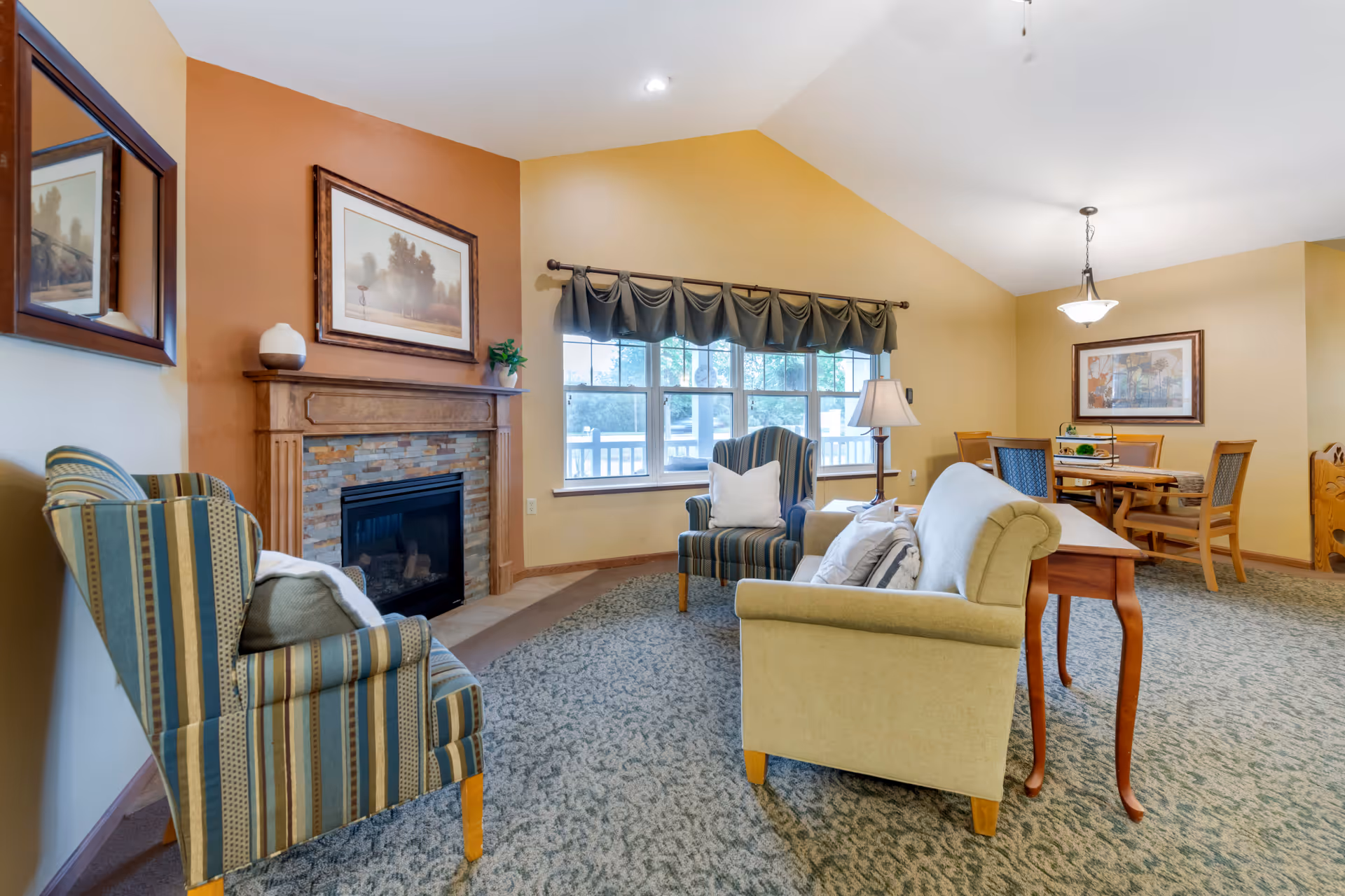 A cozy living room area with a fireplace on the left wall, two striped armchairs, a beige sofa, a wooden side table with a lamp, and a window with a dark valance. In the background, there is a dining area with a wooden table and chairs, a hanging light fixture, and framed artwork on the wall.