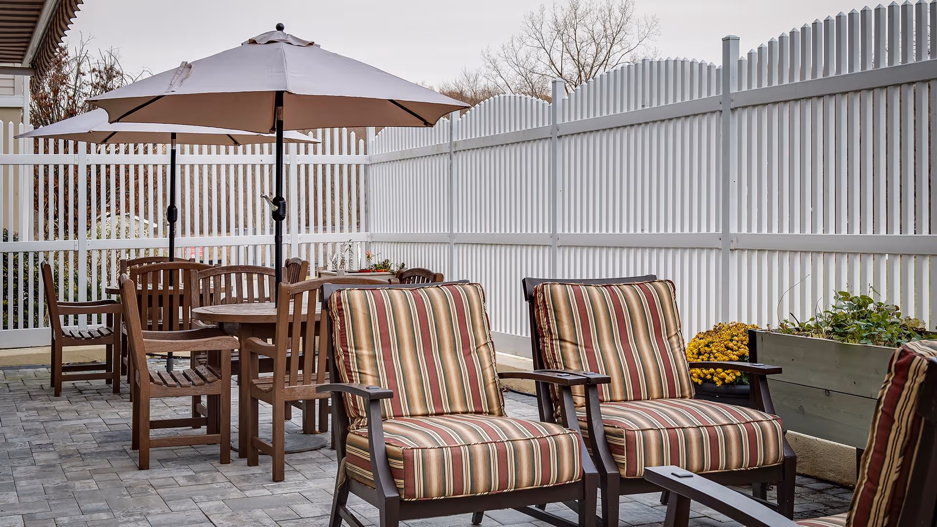 Outdoor patio with striped cushioned chairs, wooden tables and umbrellas beside a tall white fence.