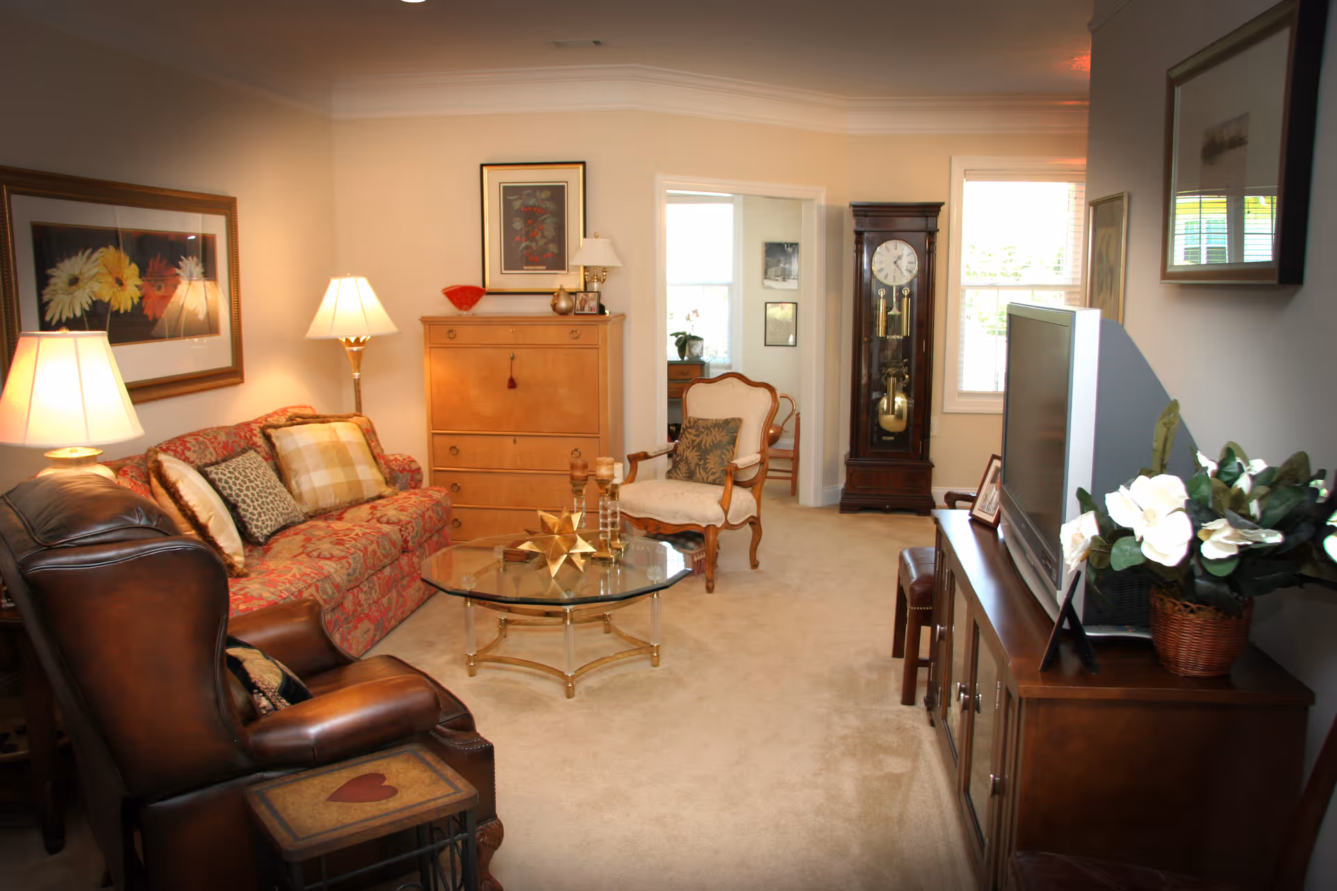 A cozy living room with a floral patterned sofa, a leather armchair, a glass coffee table with decorative items, a wooden cabinet with a flat-screen TV, a grandfather clock, and framed artwork on the walls. The room is softly lit by table lamps and natural light from windows.
