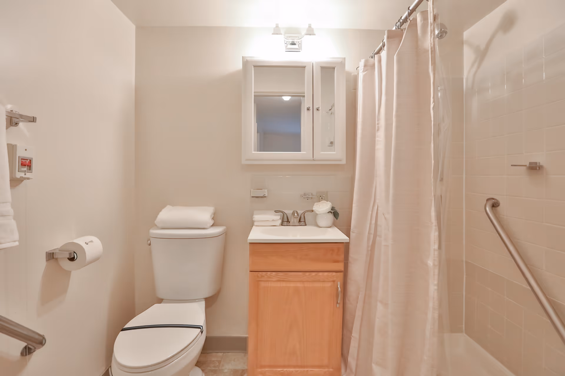 A clean and well-lit bathroom featuring a toilet with folded towels on top, a wooden vanity with a white sink and faucet, a mirrored medicine cabinet above the sink, and a shower area with a beige curtain and grab bars for safety.