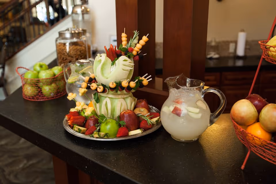 A countertop display featuring a carved melon centerpiece with fruit skewers, surrounded by various fresh fruits including apples, strawberries, and pears. There is also a glass pitcher filled with a fruit-infused beverage and a basket of green apples in the background.