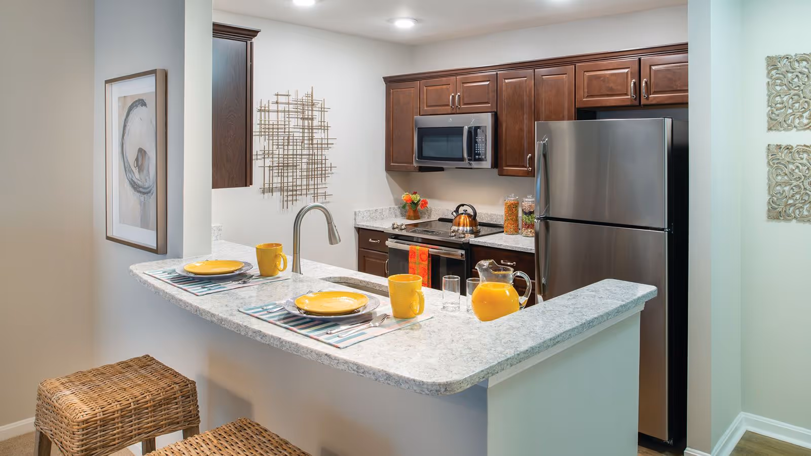 A modern kitchen with dark wood cabinets, stainless steel refrigerator and microwave, and a marble countertop breakfast bar set with yellow plates, mugs, and a pitcher of orange juice. Two wicker stools are placed at the breakfast bar, and decorative wall art is visible on the walls.