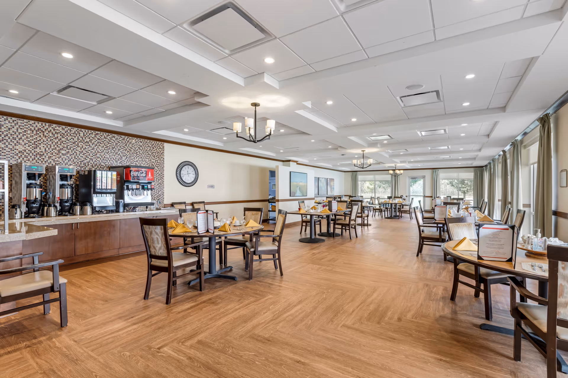 Spacious dining room in a senior living facility with multiple tables set with yellow napkins, menus, and chairs. The room features large windows with curtains allowing natural light, a beverage station with coffee and soda machines, wooden flooring, and ceiling lights including chandeliers.