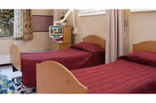 Two twin beds with burgundy bedspreads in a shared senior facility bedroom with a small TV, window, and privacy curtain.