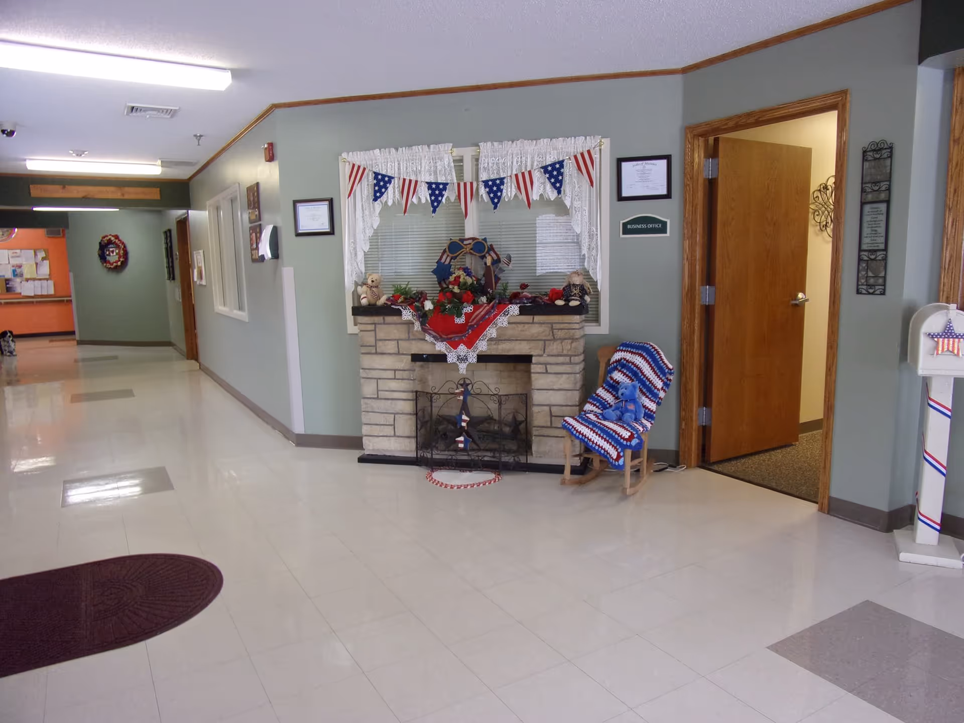 A clean and well-lit hallway in a nursing home with a decorative stone fireplace adorned with patriotic bunting, lace curtains, and various decorations. To the right of the fireplace is a wooden rocking chair covered with a red, white, and blue crocheted blanket and a blue teddy bear. An open door labeled 'Business Office' is visible, along with a white mailbox decorated with patriotic colors. The hallway extends to the left with bulletin boards and a wreath on the wall.