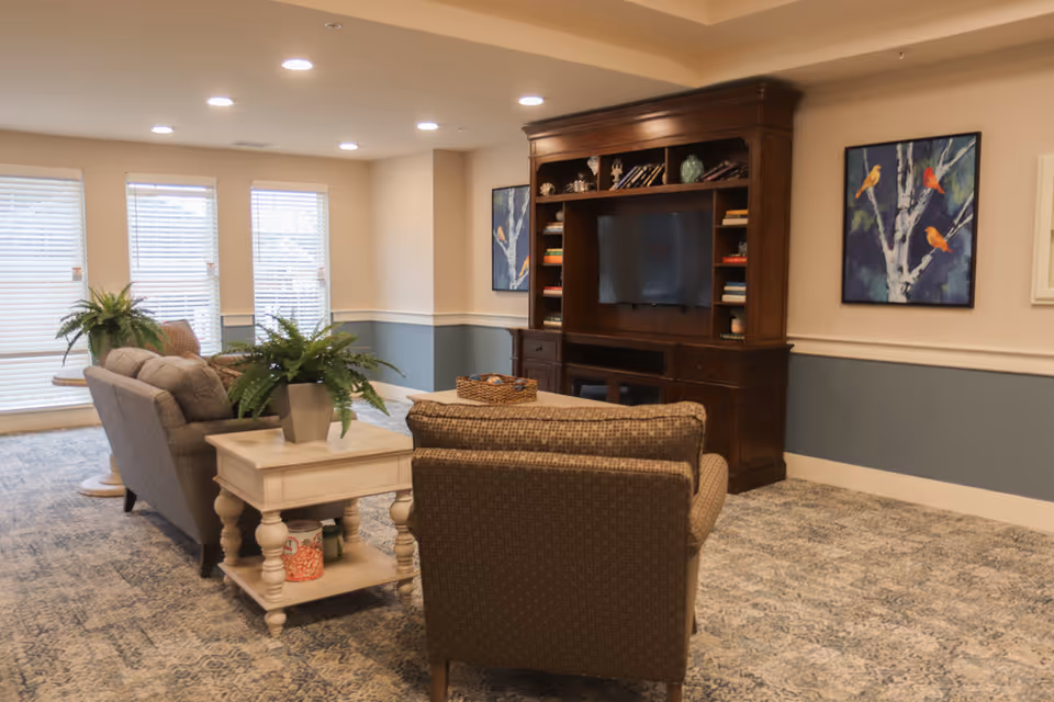 A cozy living room area with a patterned carpet, two armchairs, a sofa, and a wooden entertainment center with a TV and shelves filled with books and decorative items. There are two framed paintings of birds on the walls and potted plants on a side table and a round table near large windows with blinds.