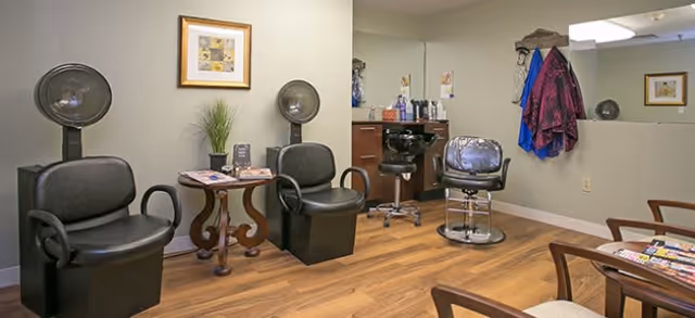 Interior view of a senior community hair salon with two black hair drying chairs, a styling chair, a small table with magazines and a plant, a mirror on the wall, and hair care products on a counter.