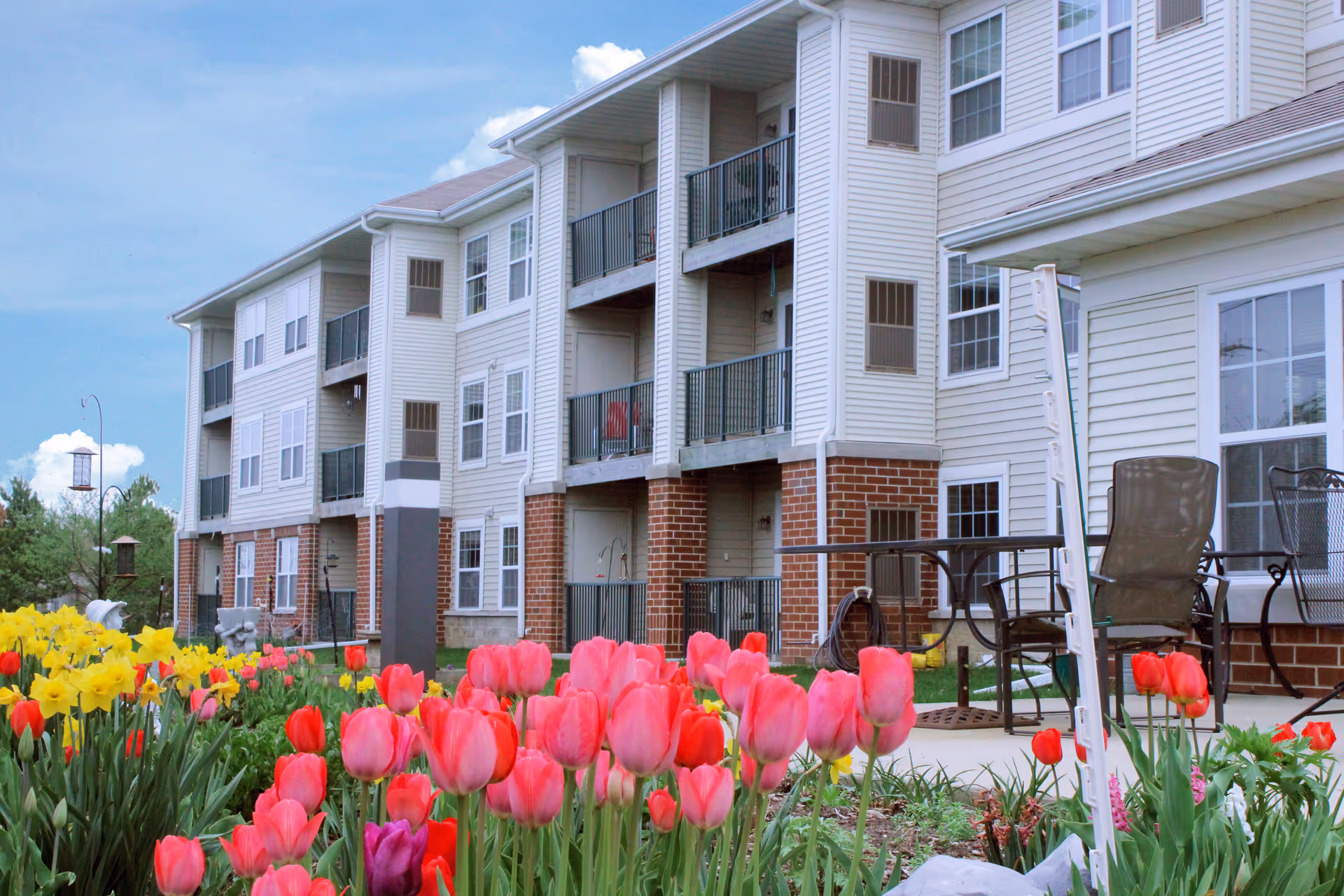 Exterior view of a multi-story residential building with balconies and a garden in the foreground featuring vibrant pink, red, and yellow tulips under a partly cloudy blue sky.