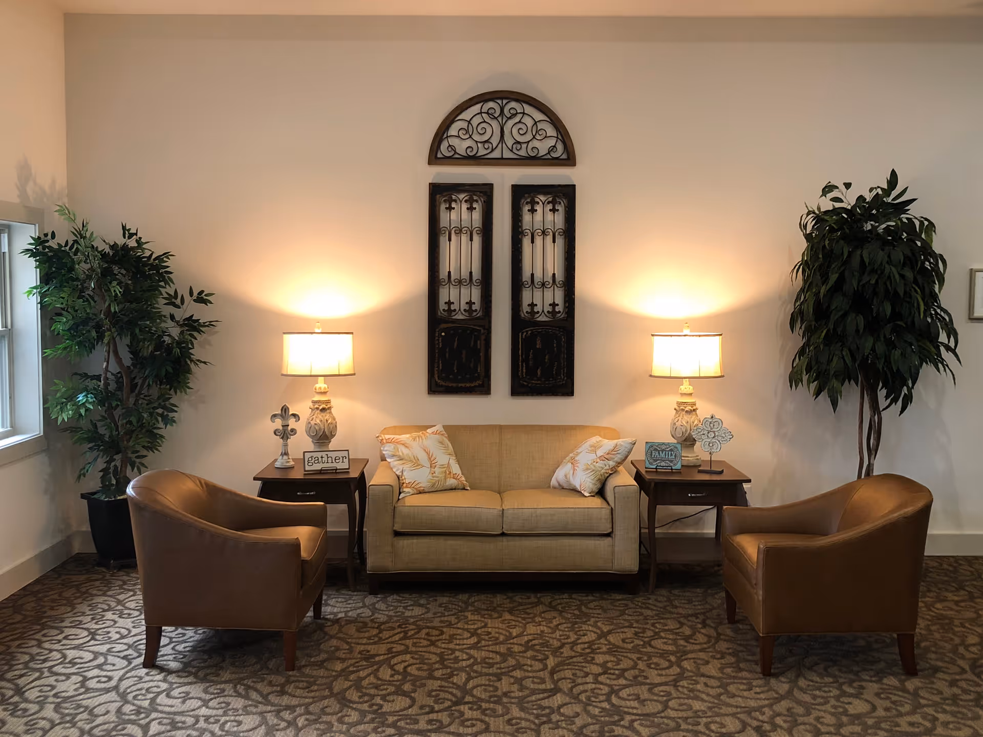 A cozy seating area with a beige loveseat adorned with two patterned pillows, flanked by two wooden side tables each holding a lamp and decorative items. Two brown leather armchairs face the loveseat. The wall behind features decorative wrought iron art pieces and two large potted plants are placed on either side of the seating arrangement.
