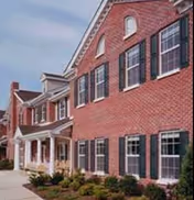 Exterior front facade of a red brick, multi-story senior living building with white trim, black shutters, a covered porch, and a landscaped walkway.