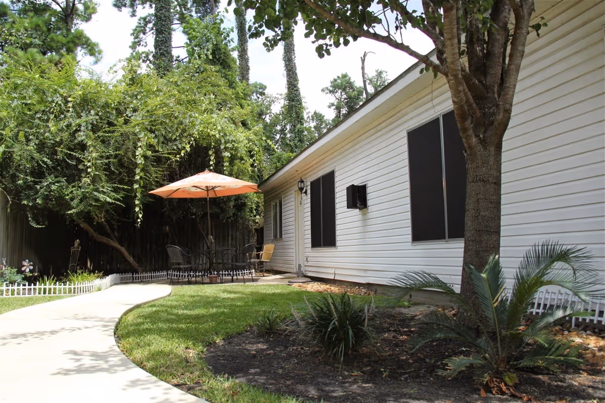 Side view of a white cottage with a curved walkway, lawn, patio table with umbrella, and surrounding trees.