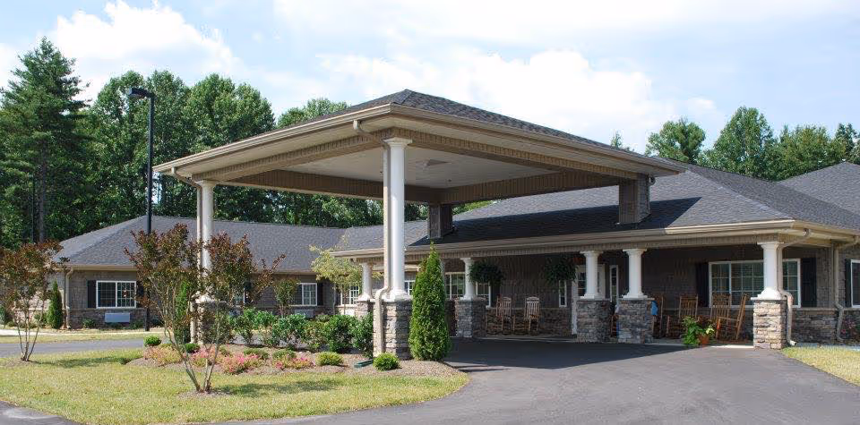 Exterior view of Mitchell House senior living facility showing a covered entrance with white columns, stone accents, rocking chairs on the porch, landscaped greenery, and a paved driveway under a partly cloudy sky.