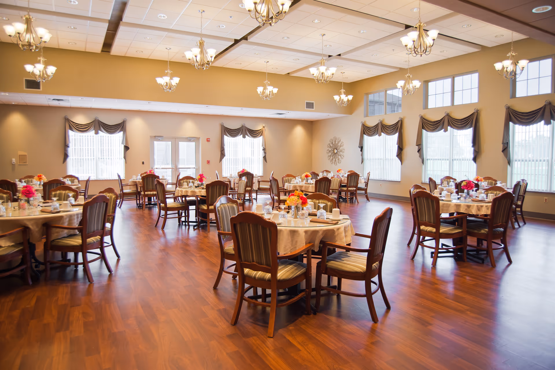 A spacious dining room with multiple round tables covered with beige tablecloths, each set with glassware, cups, and floral centerpieces. The room has large windows with draped curtains, wooden flooring, and several elegant chandeliers hanging from the ceiling.