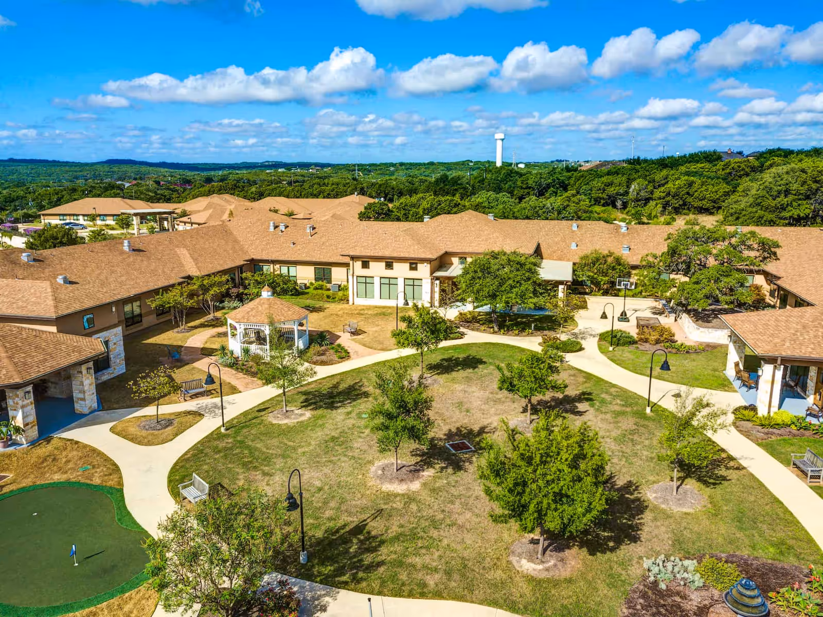 Aerial view of The Auberge at Bee Cave senior living facility showing a courtyard with walking paths, small trees, benches, a white gazebo, and a putting green. The buildings surrounding the courtyard have tan roofs and beige walls, with a backdrop of green hills and a blue sky with scattered clouds.