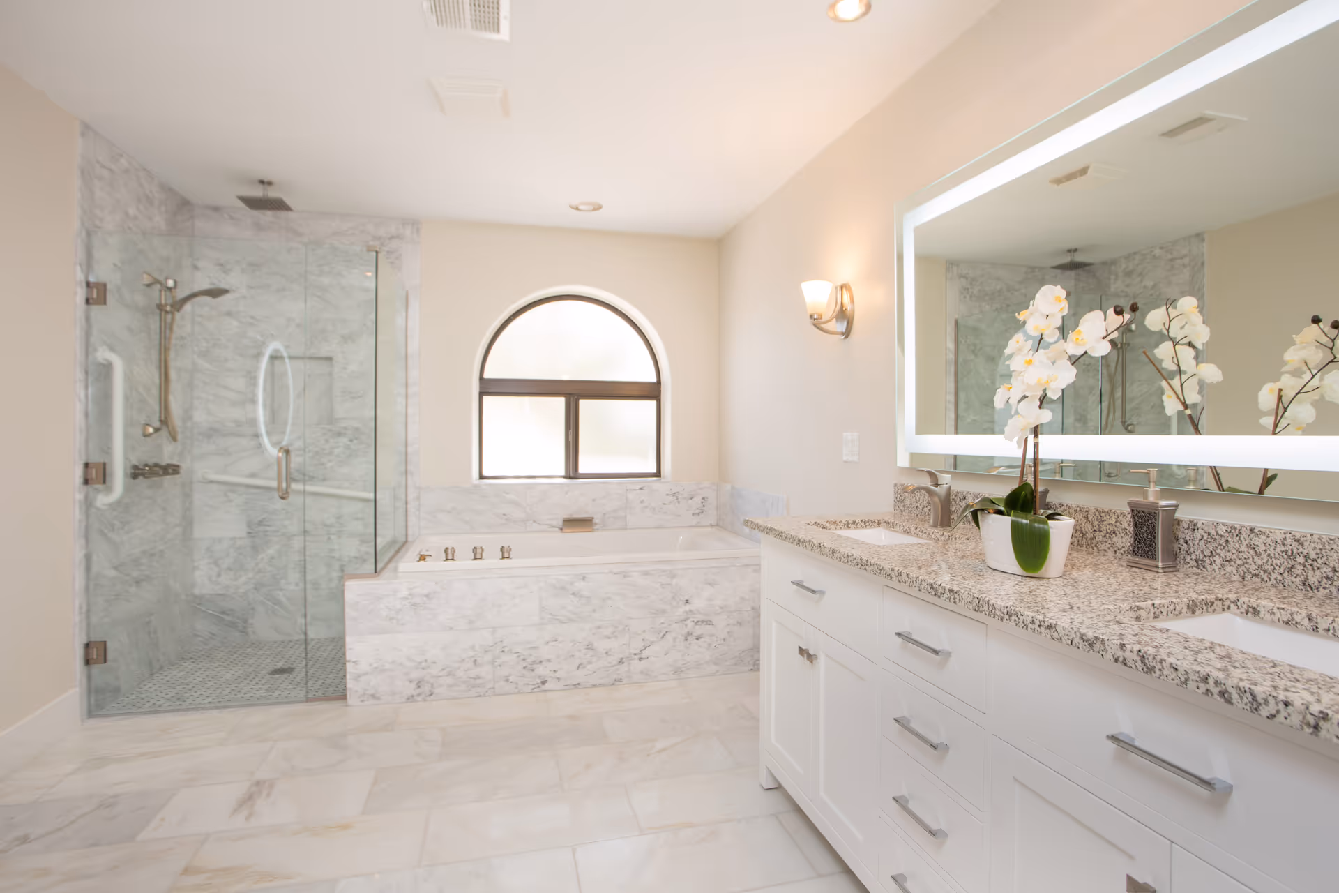 Bright and spacious bathroom with a large marble-tiled walk-in shower with glass doors, a built-in bathtub with marble surround beneath a frosted arched window, and a double sink vanity with granite countertop and a large illuminated mirror. A white orchid plant and soap dispenser are placed on the vanity.