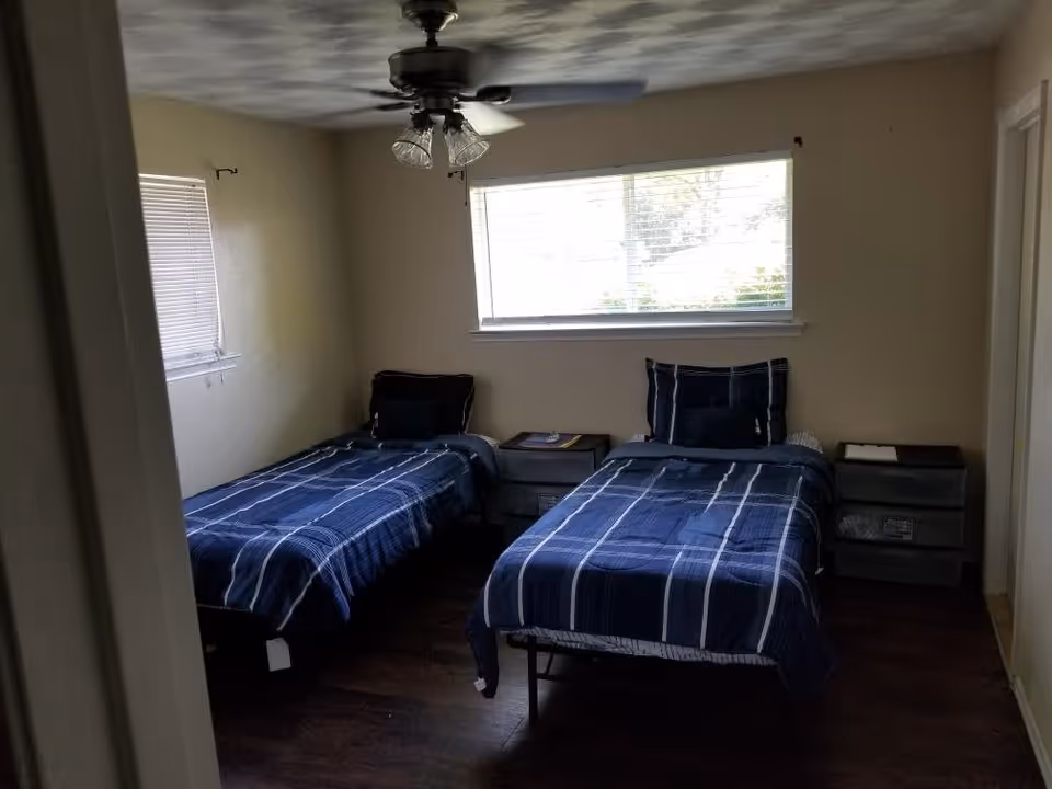 A bedroom with two single beds covered in blue plaid bedding, each with a pillow. There are two small nightstands between and beside the beds. A window with blinds is above the beds, and a ceiling fan with light fixtures is mounted on the ceiling.