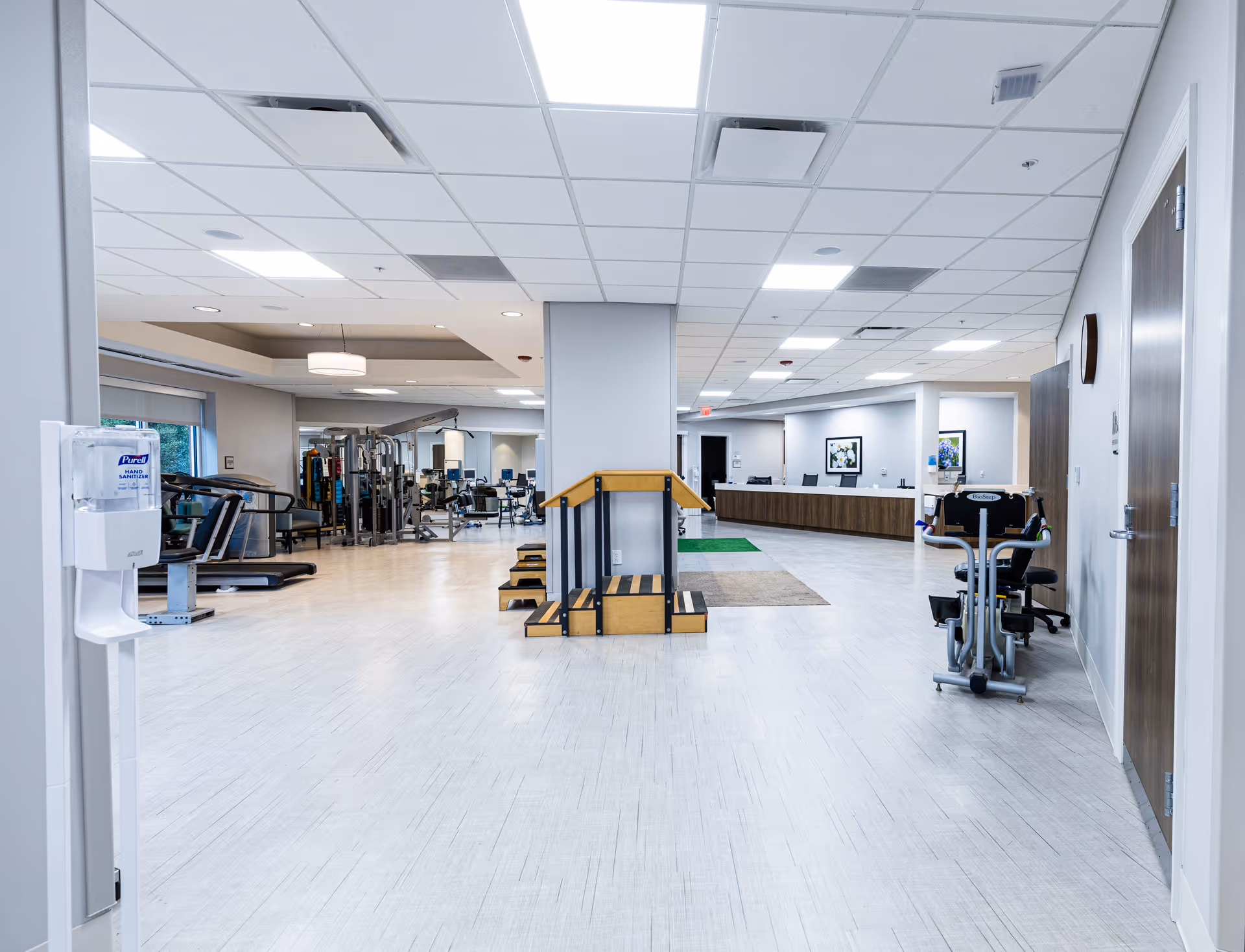 A spacious rehabilitation or physical therapy room with exercise equipment including treadmills, weight machines, and a set of wooden stairs with handrails. The room has bright overhead lighting, light-colored flooring, and a hand sanitizer dispenser mounted on the left wall near the entrance. In the background, there is a reception desk with chairs and framed artwork on the walls.