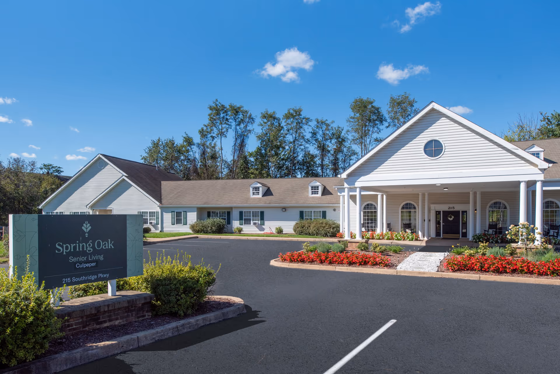 Exterior view of Spring Oak Senior Living facility in Culpeper, showing a white building with a covered entrance, surrounded by landscaped flower beds and greenery under a clear blue sky.