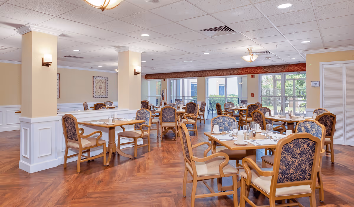 A bright and spacious dining room with multiple wooden tables and cushioned chairs arranged neatly. Each table is set with white napkins, glasses, and silverware. Large windows in the background provide natural light and a view of greenery outside. The room features warm yellow walls, white wainscoting, and wooden flooring.