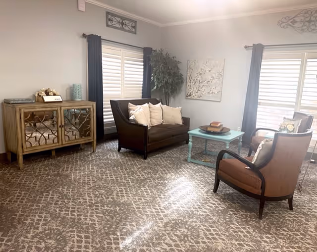 Sunlit sitting room with a sofa, two armchairs, a teal coffee table, sideboard and patterned carpet.