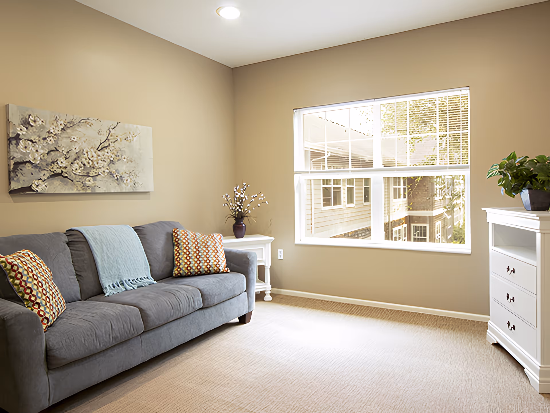 A cozy living room with a gray couch adorned with two patterned pillows and a light blue throw blanket. There is a white side table with a vase of flowers next to the couch, a large window letting in natural light, and a white chest of drawers with a green plant on top. A floral painting hangs on the beige wall above the couch.