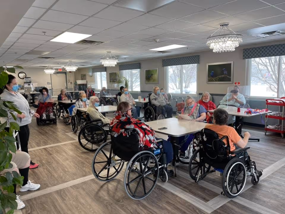A group of elderly individuals, many in wheelchairs, seated around tables in a spacious, well-lit room with large windows. Several people are wearing face masks, and a staff member stands to the left. The room has wood flooring, modern light fixtures, and framed pictures on the walls.