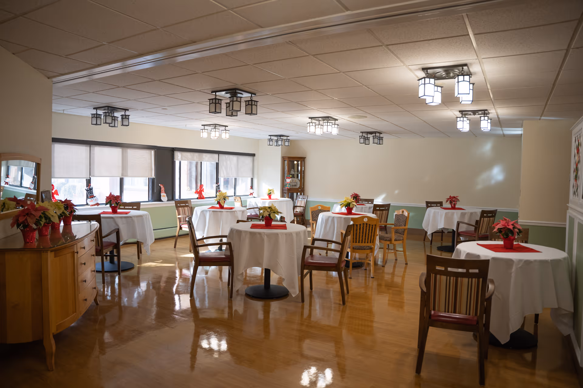 A dining room with several round tables covered with white tablecloths, each decorated with a small red or yellow poinsettia centerpiece. Wooden chairs surround the tables. The room has large windows with blinds partially drawn, letting in natural light. The floor is polished wood, and the ceiling has multiple modern light fixtures. A wooden sideboard with additional poinsettias is visible on the left side of the room.