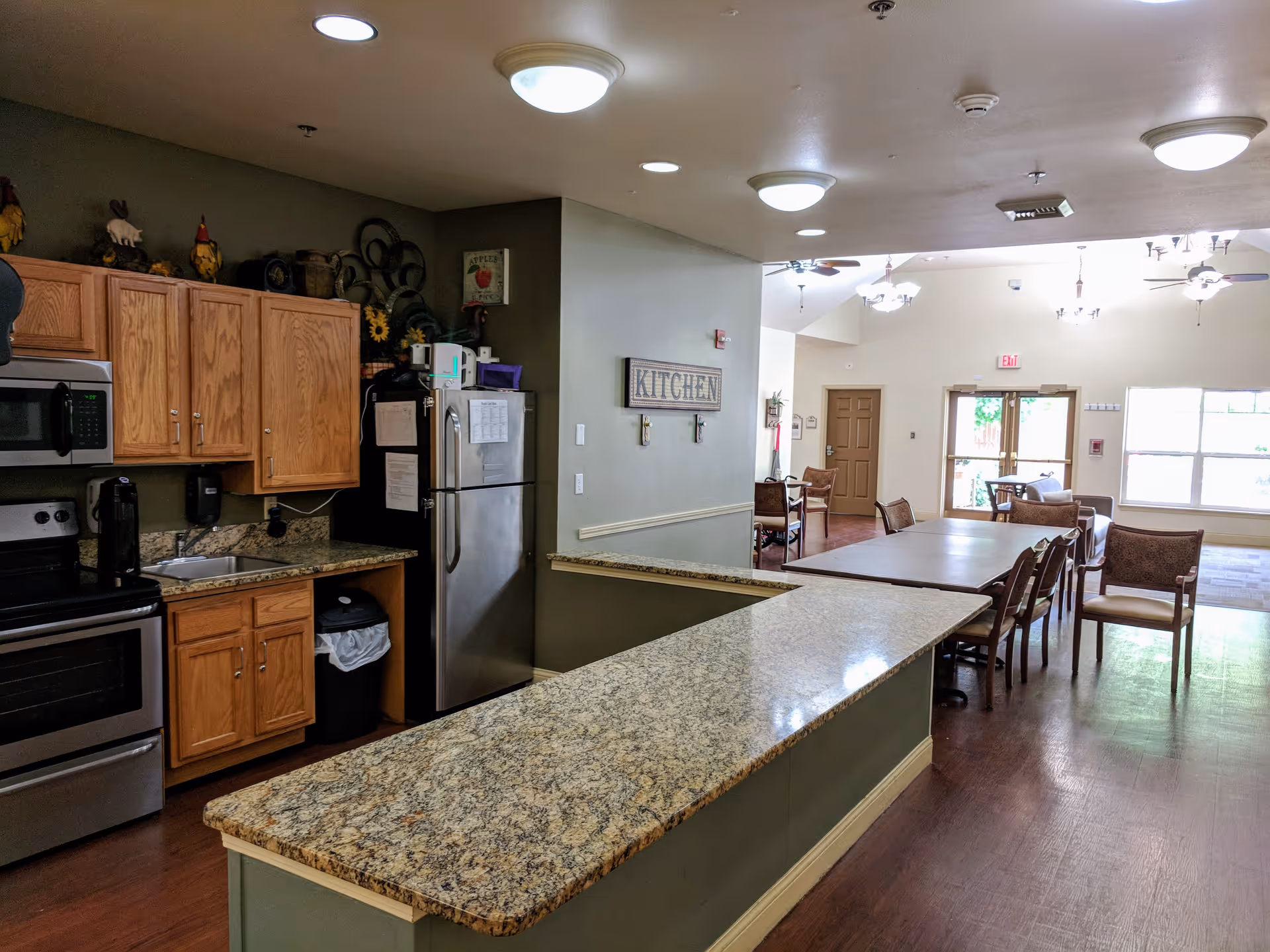 Interior view of a senior living facility kitchen and dining area. The kitchen features wooden cabinets, a stainless steel refrigerator, microwave, and stove. A granite countertop extends into a dining area with tables and chairs. The space is well-lit with ceiling lights and natural light from large windows in the background.