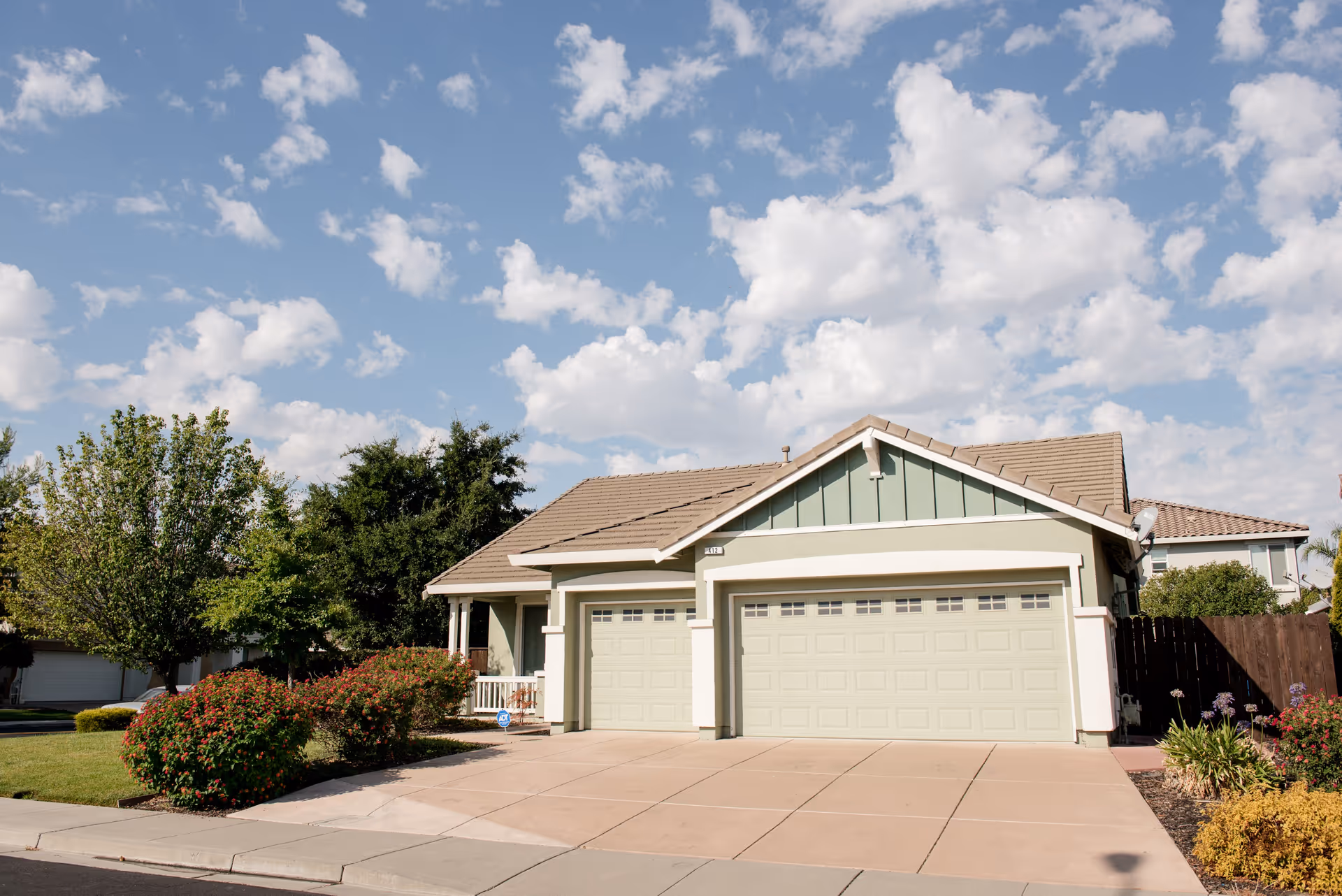 A single-story residential house with a three-car garage, light green exterior walls, and a beige tiled roof. The driveway is spacious and clean, with well-maintained bushes and trees in the front yard under a partly cloudy blue sky.
