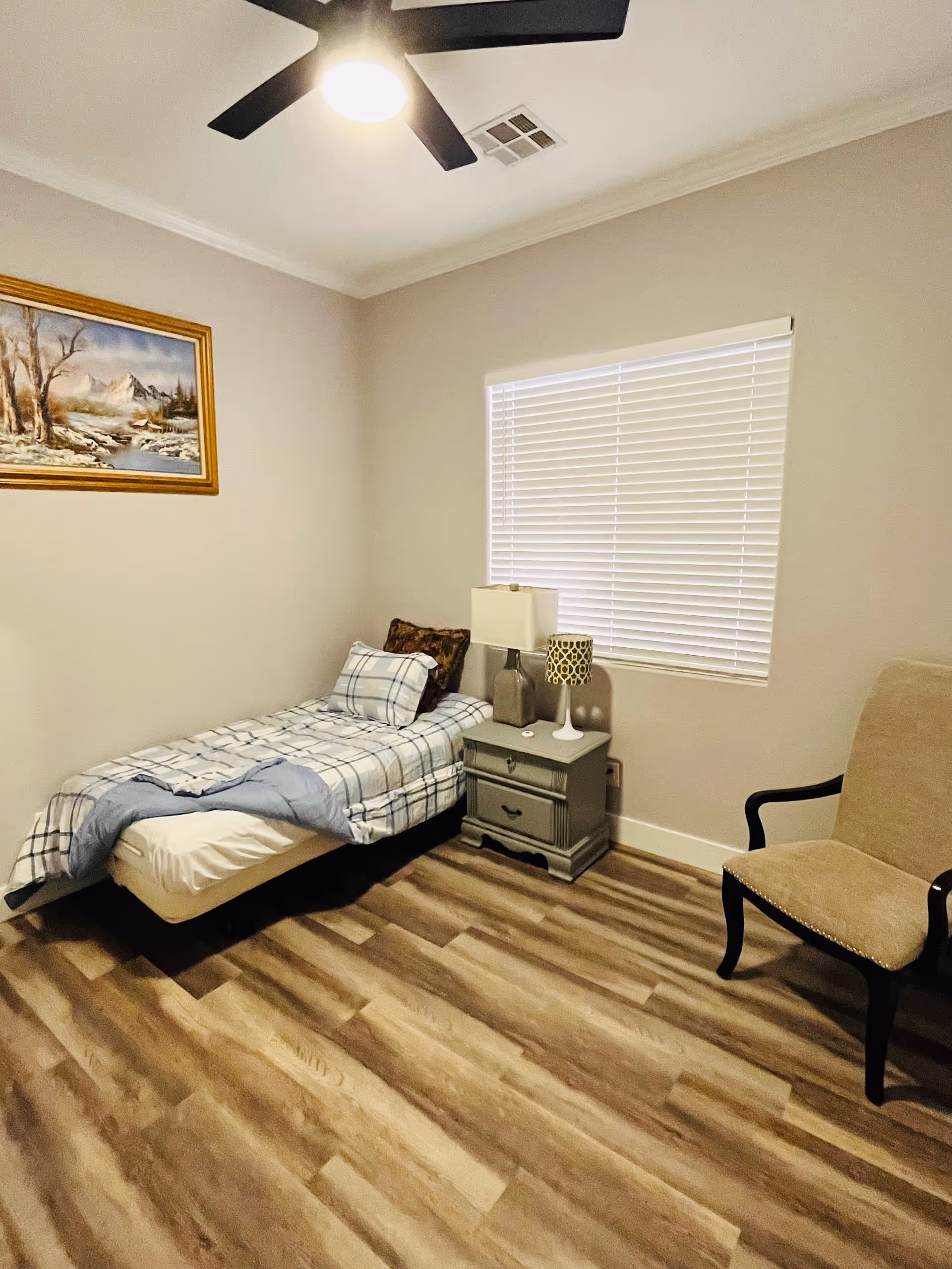 A small bedroom with a single bed covered in plaid bedding, a nightstand with two lamps, a beige upholstered chair, a window with closed blinds, a ceiling fan with a light, and a framed landscape painting on the wall.