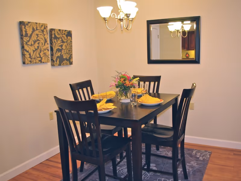 A dining room with a dark wooden table set for four with yellow napkins and plates. There is a floral centerpiece on the table. Four matching dark wooden chairs surround the table. The room has light-colored walls with two decorative wall hangings on one side and a large mirror on another wall. A chandelier with three lights hangs above the table. The floor is wooden with a patterned rug under the table.