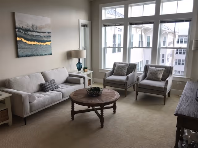 Bright living room with a sofa, two armchairs, a round wooden coffee table, large windows, and wall art.